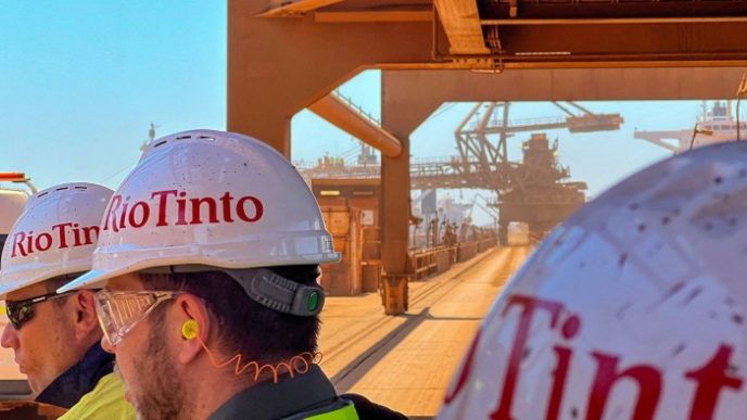 Visitors to Rio Tinto’s iron ore export port of Dampier walk in front of a loader transferring iron ore into a bulk carrier