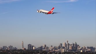 A white and red Qantas plane in the blue sky over a city skyline