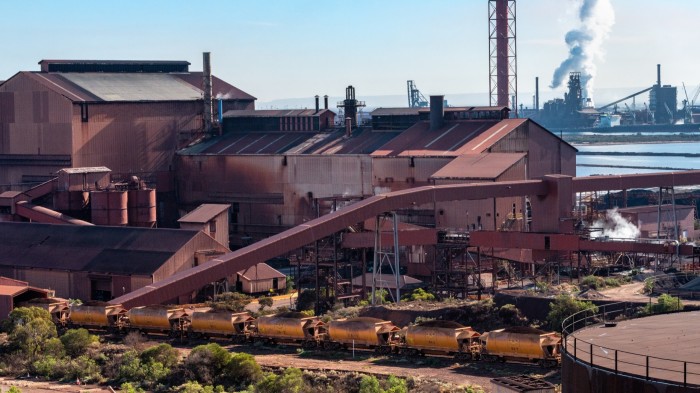 The Whyalla steelworks, featuring large industrial buildings with metal roofs and chimneys emitting smoke. In the foreground, there are freight train cars on tracks, and vegetation is visible around the site.