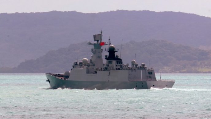 A frigate travels in turquoise waters against a backdrop of hilly terrain under a clear sky