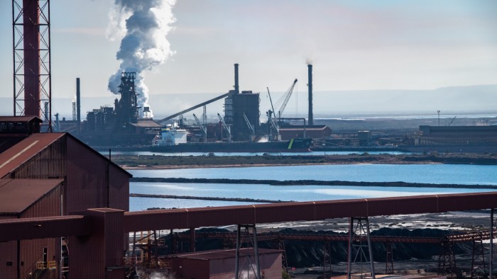 The Whyalla steelworks, featuring industrial buildings and large chimneys emitting smoke. A body of water is visible in the foreground, with cranes and shipping vessels nearby.