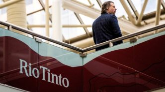 A man stands on an upper-level walkway, partially turned away from the camera, near a large Rio Tinto sign