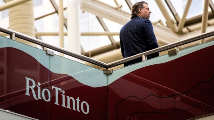 A man stands on an upper-level walkway, partially turned away from the camera, near a large Rio Tinto sign