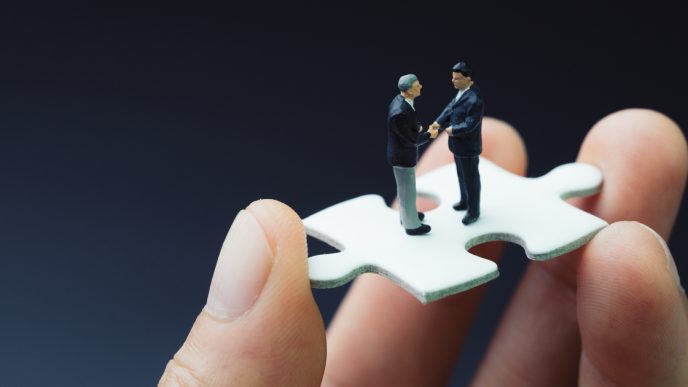 Hand holding puzzle piece with two small male figures on top shaking hands